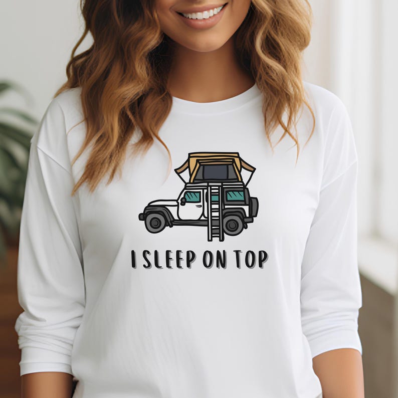 May include: White long-sleeve t-shirt with a cartoon illustration of a car with a rooftop tent and a ladder. The text "I SLEEP ON TOP" is printed below the image.