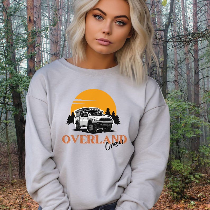 May include: A white sweatshirt featuring a graphic of a white truck with a roof rack and a yellow sun behind it. The text "OVERLAND Crew" is printed below the truck.