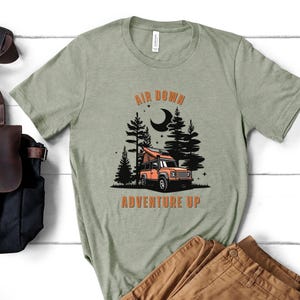 May include: A green t-shirt with the text "Air Down Adventure Up" in orange lettering. The design features a cartoon illustration of a camper van in a forest setting with a crescent moon in the sky.