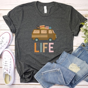 May include: Dark gray t-shirt featuring a brown camper van graphic with luggage on top and the word "LIFE" in colorful letters. The shirt is paired with white sneakers and blue jeans.