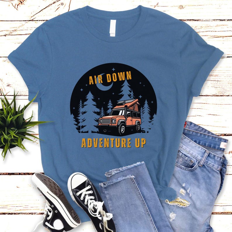 Puede incluir: Una camiseta azul con un gr&aacute;fico de un coche en un bosque con el texto "Air Down Adventure Up".