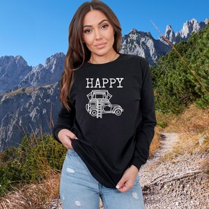 May include: A black long-sleeve shirt with the text "HAPPY" and a white illustration of a car with a rooftop tent. The person wearing the shirt is standing in front of a mountain background.