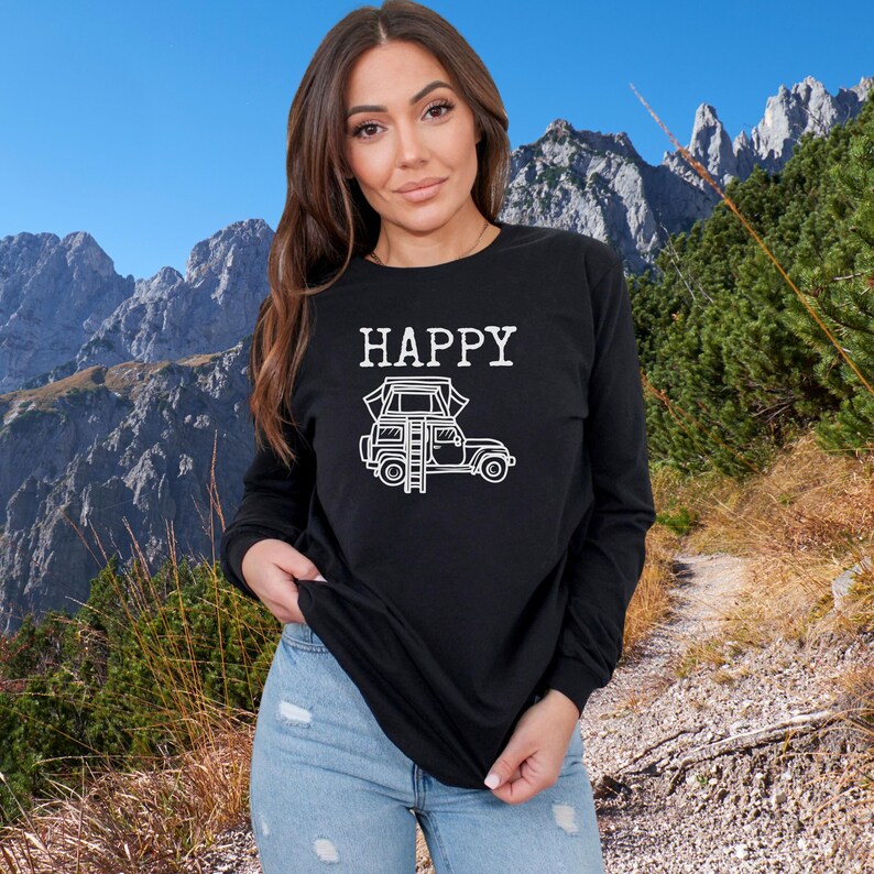 May include: A black long-sleeve shirt with the text "HAPPY" and a white illustration of a car with a rooftop tent. The person wearing the shirt is standing in front of a mountain background.