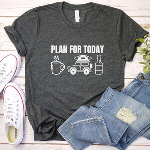 Puede incluir: Camiseta gris oscuro con el texto blanco "PLAN FOR TODAY" e ilustraciones de una taza de caf&eacute;, una furgoneta camper y una botella. Zapatillas blancas y vaqueros azules junto a la camiseta.