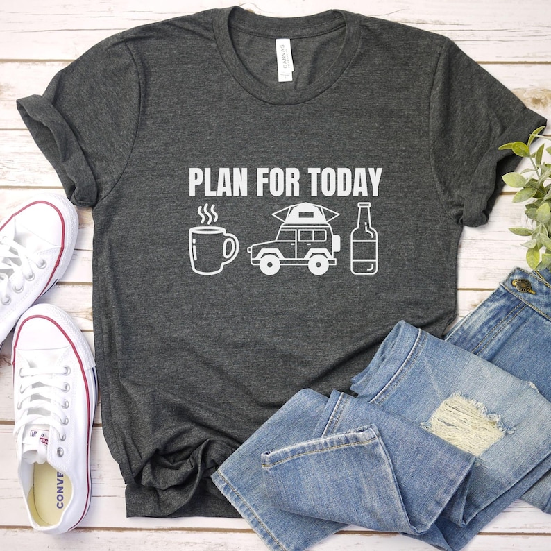Puede incluir: Camiseta gris oscuro con el texto blanco "PLAN FOR TODAY" e ilustraciones de una taza de caf&eacute;, una furgoneta camper y una botella. Zapatillas blancas y vaqueros azules junto a la camiseta.