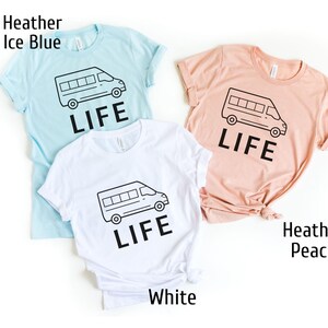 Op de afbeelding: Drie T-shirts met een afbeelding van een busje met het woord "LIFE" erop gedrukt. De T-shirts zijn in drie kleuren: heide-ijsblauw, wit en heideperzik.