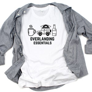Puede incluir: Camiseta blanca con un gr&aacute;fico negro de un coche, una taza de caf&eacute; y una botella. El texto "Overlanding Essentials" est&aacute; impreso debajo del gr&aacute;fico.