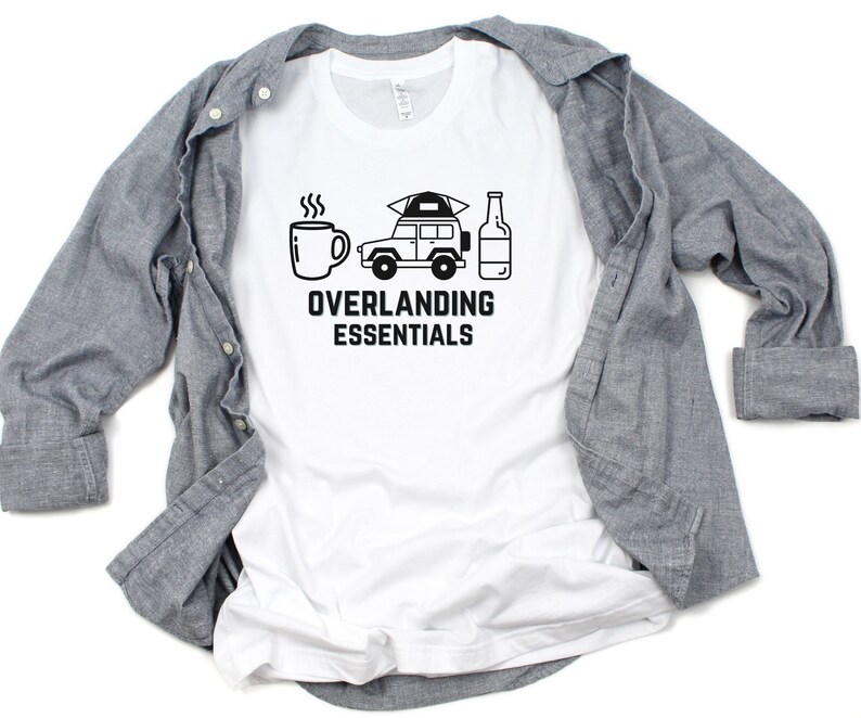 Puede incluir: Camiseta blanca con un gr&aacute;fico negro de un coche, una taza de caf&eacute; y una botella. El texto "Overlanding Essentials" est&aacute; impreso debajo del gr&aacute;fico.