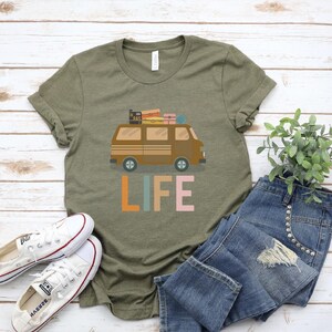 May include: Olive green t-shirt featuring a brown van with luggage on top and the word "LIFE" in colorful letters. The shirt is paired with blue jeans and white sneakers.