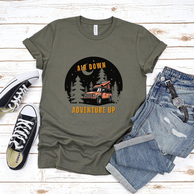 Puede incluir: Una camiseta verde con un gr&aacute;fico de una furgoneta de camping en un bosque por la noche. El texto "Air Down Adventure Up" est&aacute; impreso en la camiseta.