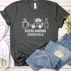 Puede incluir: Una camiseta gris oscuro con texto blanco que dice "Overlanding Essentials". El gr&aacute;fico en la camiseta incluye una taza de caf&eacute;, un jeep con una tienda de campa&ntilde;a en la parte superior y una botella de cerveza.
