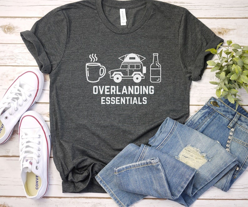 Puede incluir: Una camiseta gris oscuro con texto blanco que dice "Overlanding Essentials". El gr&aacute;fico en la camiseta incluye una taza de caf&eacute;, un jeep con una tienda de campa&ntilde;a en la parte superior y una botella de cerveza.