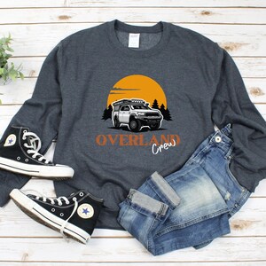 May include: A dark grey sweatshirt featuring a white truck graphic, a sunset, and the text "Overland Crew" in orange. The sweatshirt is laid out on a wooden surface with a pair of black trainers and blue jeans.