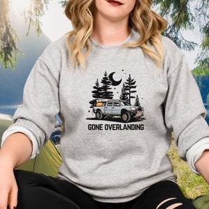 May include: A gray sweatshirt with a black graphic of a truck with a rooftop tent driving through a forest at night. The text "Gone Overlanding" is printed below the truck.