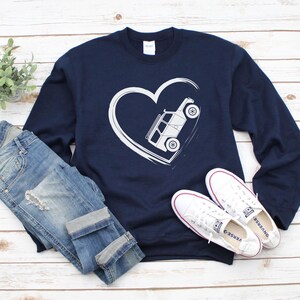May include: Navy blue sweatshirt with a white heart design containing a vehicle. The sweatshirt is paired with blue jeans and white sneakers. The design features a white vehicle inside a white heart.