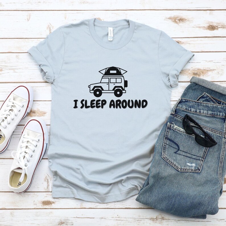 Pu&ograve; includere: Una maglietta blu chiaro con una grafica nera di un'auto con una tenda sul tetto. Il testo "I SLEEP AROUND" &egrave; stampato sotto l'auto.