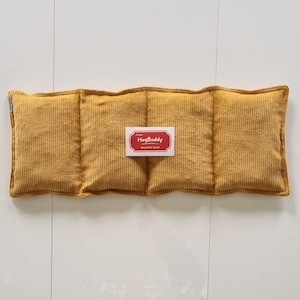Puede incluir: Paquete térmico de pana amarillo mostaza con forma rectangular. El paquete está dividido en cuatro secciones y tiene una etiqueta que dice "HugBuddy" en un diseño rojo y blanco. El paquete térmico está diseñado para una calidez calmante.