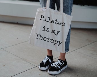 Può includere: Una borsa tote color crema con la scritta "Pilates is my Therapy" in nero. La borsa è tenuta da una persona che indossa scarpe da ginnastica nere con lacci bianchi e jeans blu.