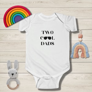 Pode incluir: Macacão branco para bebê com texto preto que diz "TWO COOL DADS" com um par de óculos de sol no lugar do "O" em "COOL".