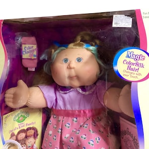 May include: A Cabbage Patch Kids doll in a box. The doll has blue eyes, blonde hair with blue streaks, and is wearing a purple dress with a pink floral skirt. The box includes a color changer and a book titled "One of a Kind Family!"