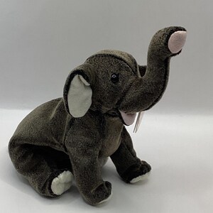 May include: A plush elephant toy in shades of gray, with white accents on the ears and feet, and a pink trunk tip. The elephant is in a seated position with its trunk raised. A small tag is attached.