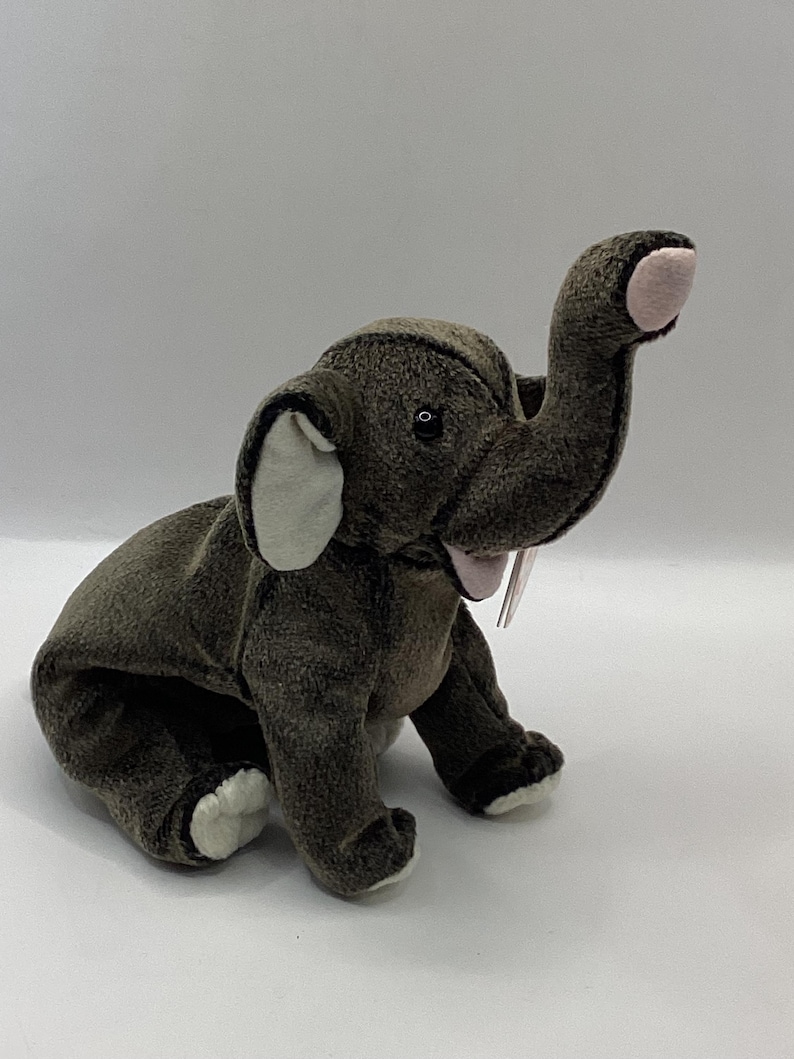 May include: A plush elephant toy in shades of gray, with white accents on the ears and feet, and a pink trunk tip. The elephant is in a seated position with its trunk raised. A small tag is attached.