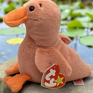 May include: A plush duck toy with soft, dusty pink fur and a bright orange beak and webbed feet. The stuffed animal features a heart-shaped red and gold Ty Beanie Baby tag attached to its side, with black plastic eyes and a stitched mouth detail.