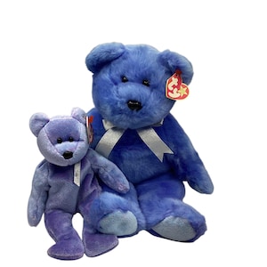 May include: Two plush teddy bears in shades of blue and purple. The larger bear is deep blue with a silver ribbon around its neck and a red Ty heart tag. The smaller bear is light purple with a silver ribbon and a red Ty heart tag.