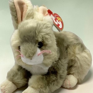 May include: A plush bunny toy in shades of gray and white, with pink accents on the inner ears, nose, and paws. The bunny has black eyes and a red tag attached to its ear. The toy is designed to look like a rabbit.