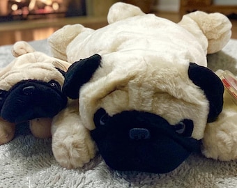 May include: Two plush pug dog toys. The larger pug is lying down, and the smaller pug is in front of it. Both have tan fur with black ears and snouts. The toys are on a light gray surface.
