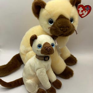 May include: Two plush Siamese cats, one larger than the other, both with brown ears and faces, blue eyes, and a cream-colored body. The larger cat has a Ty tag attached to its ear.