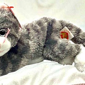 May include: A plush gray and white cat toy with a pink nose and whiskers. The stuffed animal is lying down on a white surface. A red tag is attached to the toy.