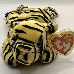 May include: A plush tiger toy with a yellow body and black stripes. The tiger has black eyes, a pink nose, and a tag that reads "Beanie Original Baby" in a heart-shaped design. The toy is made of soft, fuzzy material.