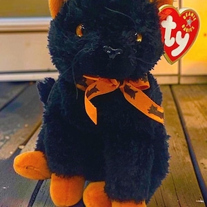 May include: A plush black cat toy with orange inner ears and orange paws. The cat has amber-colored eyes, a black nose, and whiskers. An orange ribbon with a black cat pattern is tied around its neck. A heart-shaped Ty Beanie Babies tag is attached.