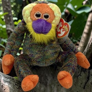May include: A plush orangutan toy with a multicolored mottled body, bright orange hands and feet, a yellow beard, and a purple nose. The face features large black eyes and a heart-shaped red tag attached to the ear with the text 2000 printed on it.