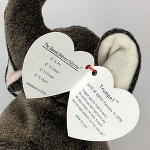 May include: A gray plush elephant toy with white and pink accents, featuring a heart-shaped tag. The tag includes the name "Trumpet", date of birth, and a poem. The toy is part of The Beanie Babies Collection.