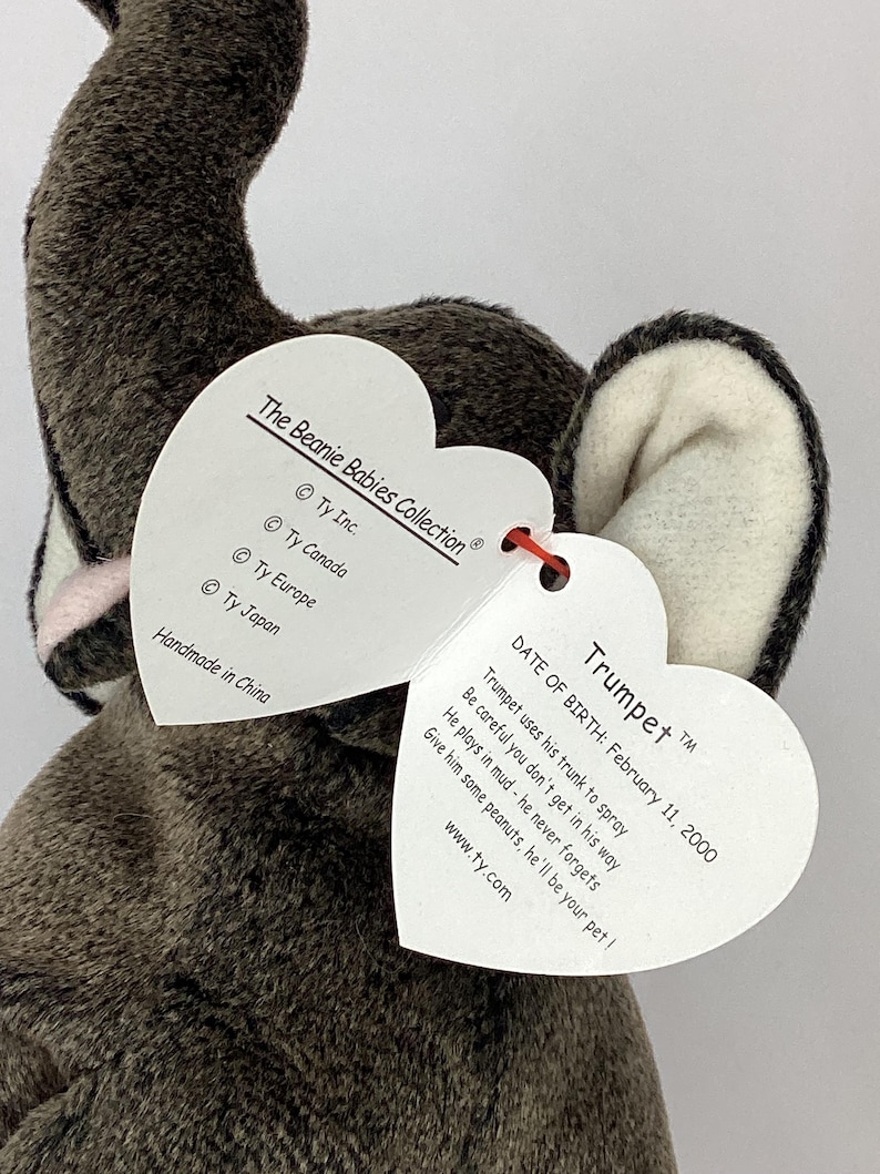 May include: A gray plush elephant toy with white and pink accents, featuring a heart-shaped tag. The tag includes the name "Trumpet", date of birth, and a poem. The toy is part of The Beanie Babies Collection.