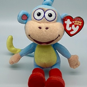 May include: A blue plush monkey toy with a tan face, large purple-rimmed eyes, and a smiling mouth. It has red boots, a yellow belly, and a yellow tail. A red heart-shaped tag with "ty" and "Original Beanie Babies" is attached.