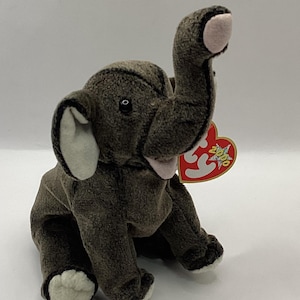 May include: A plush elephant toy in shades of brown and gray, with white accents on the ears and feet. The elephant has a raised trunk and a pink mouth. A red heart-shaped tag is attached to the ear.