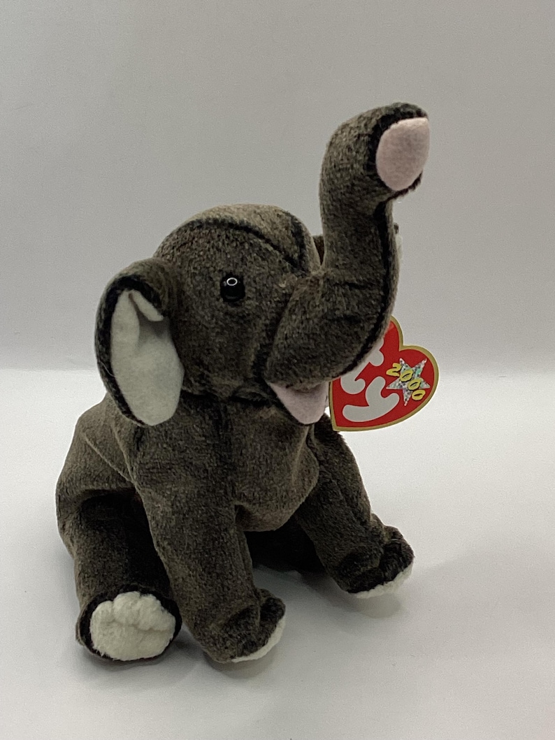 May include: A plush elephant toy in shades of brown and gray, with white accents on the ears and feet. The elephant has a raised trunk and a pink mouth. A red heart-shaped tag is attached to the ear.