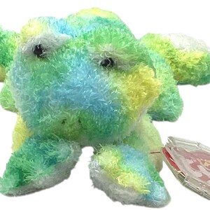 May include: A plush frog toy featuring a textured, multi-colored faux fur pattern in shades of lime green, bright yellow, and sky blue. The stuffed animal has two small black plastic eyes and a clear plastic heart-shaped tag attached to one leg.
