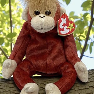 May include: A plush orangutan toy with a red body, tan fur, and white accents on the face and feet. The toy has a heart-shaped tag attached. The stuffed animal is sitting on a tree branch.
