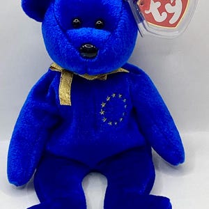 May include: A plush blue teddy bear with black eyes and nose, and a gold ribbon around its neck. The bear has a gold star design on its chest and a Ty Beanie Babies tag.