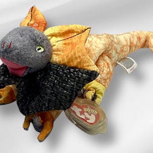 May include: A plush toy dragon with orange, yellow, and black scales. The dragon has a gray head with a pink mouth and green eyes. The toy has a Ty tag attached to it.