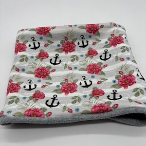 Könnte beinhalten: Ein Nackenwärmer mit maritimem Muster, verziert mit schwarzen Ankern, rosa Rosen und blauen floralen Akzenten auf einem grau-weiß gestreiften Hintergrund. Die Kanten sind mit grauem Fleece besetzt.