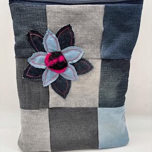 Könnte beinhalten: Eine Patchwork-Jeans-Tragetasche mit Reißverschluss. Die Tasche hat eine große, applizierte Blume mit blauen Blütenblättern, rosa Akzenten und einem schwarz-rosa Zentrum. Die Tasche besteht aus verschiedenen Blau- und Grautönen aus Jeansstoff.
