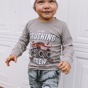 May include: A young child wearing a gray long-sleeve shirt with a red monster truck graphic and the text "Heart Crushing Crew". The child is also wearing a white beanie with a logo, distressed denim jeans, and red sneakers.