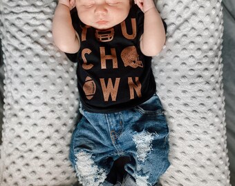 Puede incluir: Un bebé con una camiseta negra con las palabras "You  Show  Own" en letras marrones y pantalones vaqueros azules desgastados. El bebé está durmiendo en una almohada con estampado gris y blanco.