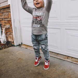 May include: A young child wearing a gray long-sleeve shirt with a red monster truck graphic and the text "Heart Crushing Crew". The child is also wearing a white beanie, blue ripped jeans, and red high-top sneakers.