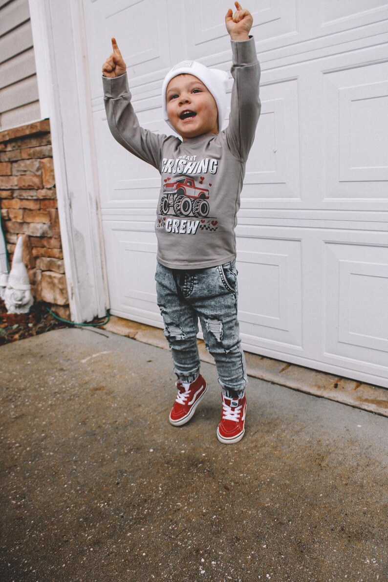 May include: A young child wearing a gray long-sleeve shirt with a red monster truck graphic and the text "Heart Crushing Crew". The child is also wearing a white beanie, blue ripped jeans, and red high-top sneakers.